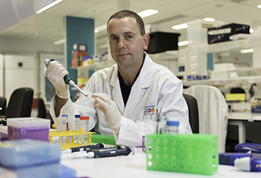 Photo of a Dr Waithman wearing a white lab coat, in a lab holding a test tube.