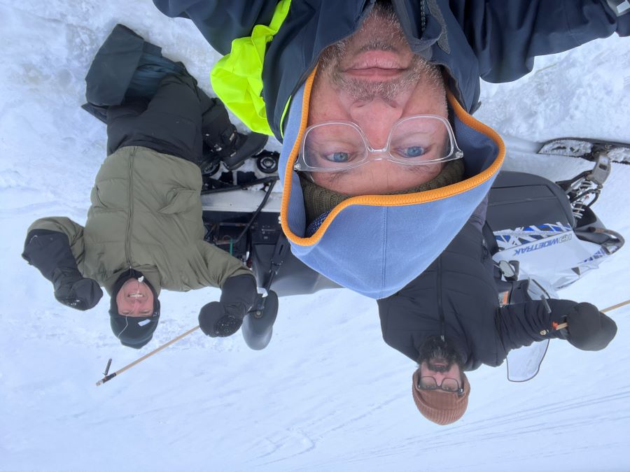 Three men ice fishing in the snow smiling and looking towards the camera