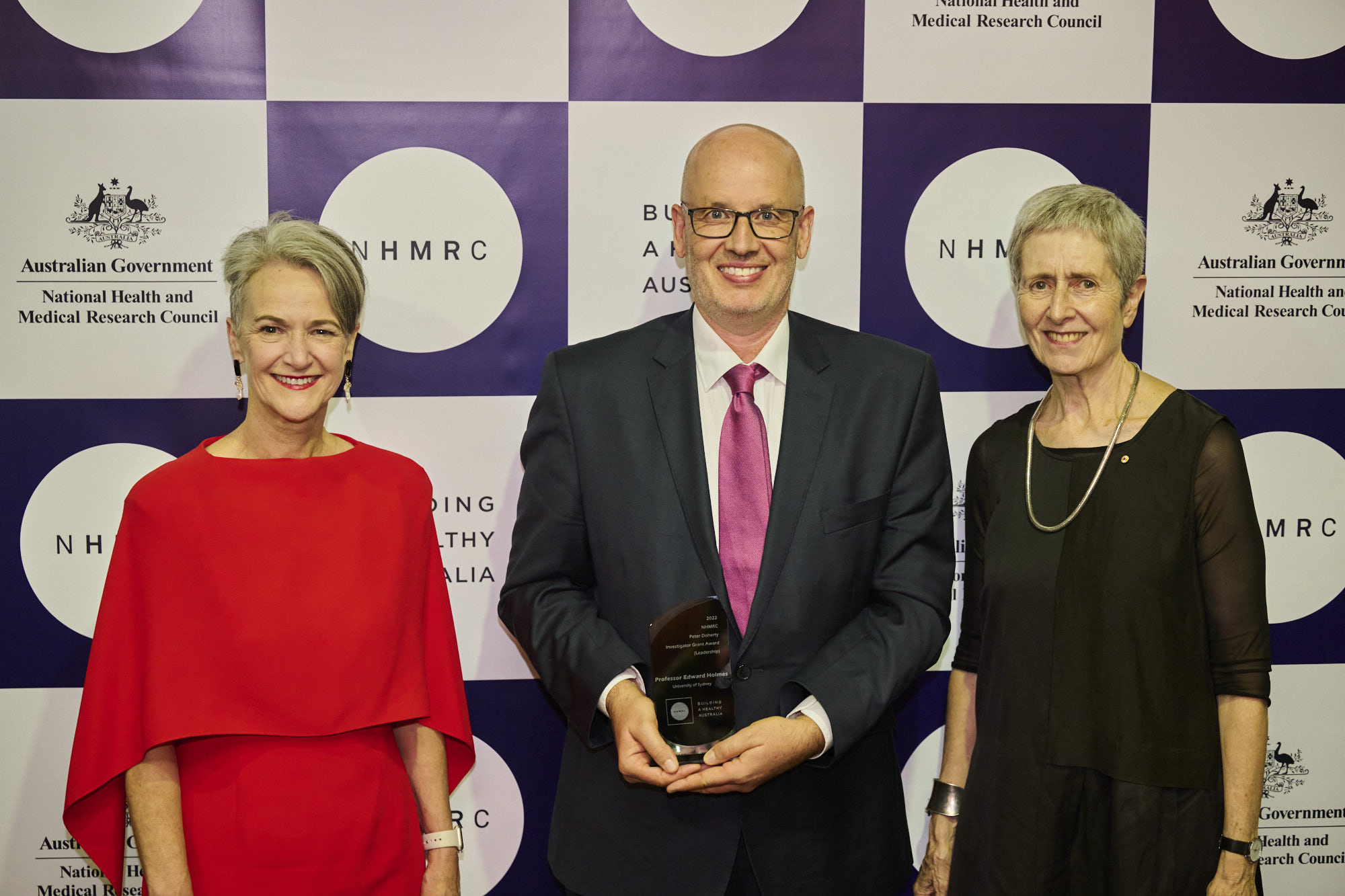 Professor Caroline Homer AO, Professor Eddie Holmes, Professor Anne Kelso AO (L-R) attending NHMRC’s Research Excellence Awards evening. Three people standing in front of media wall with the centre man holding an award.