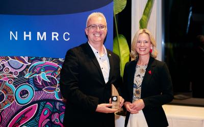Professor Arthur Christopoulos smiling at camera receiving award from Assistant Minister Rebecca White in front of NHMRC banner