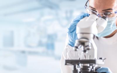 Female lab technician in protective glasses, gloves and face mask sits next to a microscope in laboratory